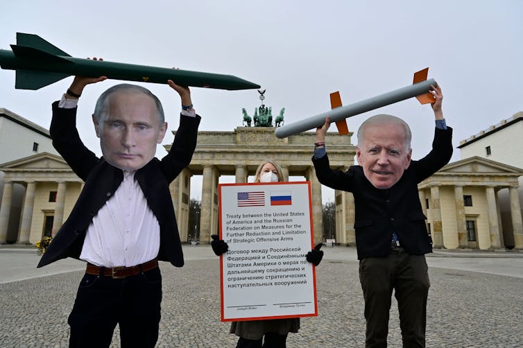 Two people wearing suits have large cut out faces of Putin and Joe Biden, They both hold fake ballistic missiles high above their heads.