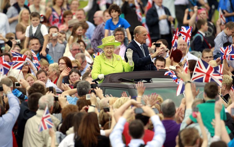 A crowd swarms a car as two people stand in through the sun roof waving.