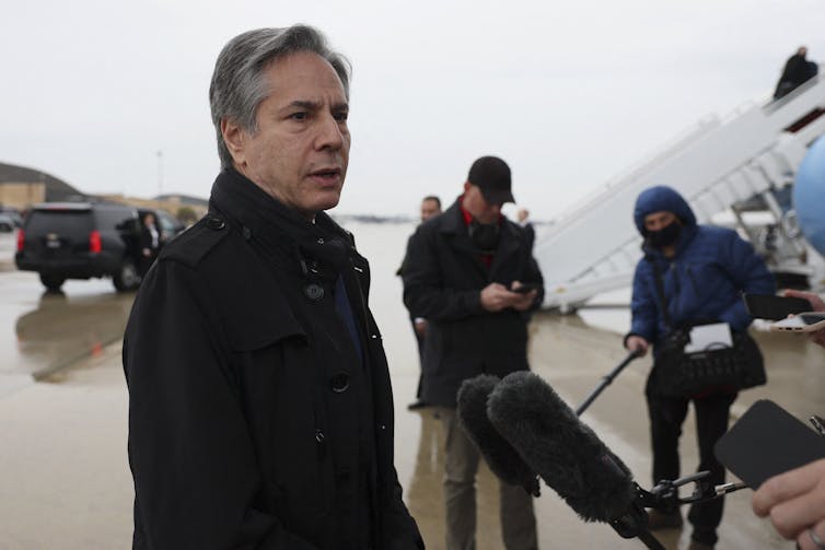 A middle-aged man with gray hair in a dark coat, talking to reporters as he boards a plane.