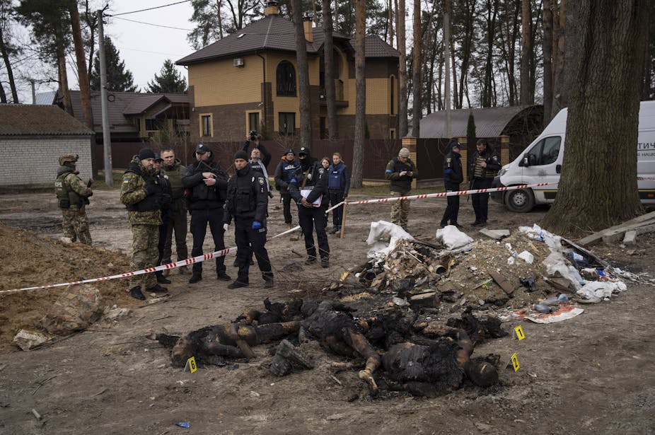 Soldiers and other official-looking people look over a pile of charred bodies on the ground.