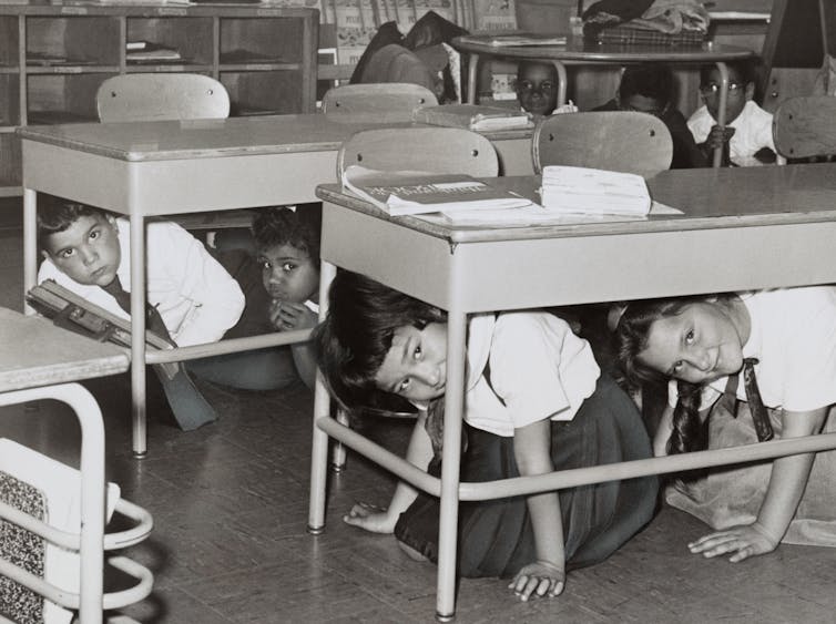 Jóvenes estudiantes se esconden bajo sus pupitres y miran hacia fuera en una foto en blanco y negro