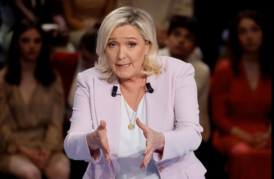 Marine Le Pen, un peu courbée, les mains tendues, parle, avec un auditoire derrière elle