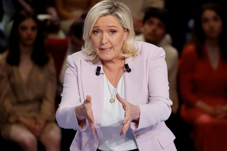 Marine Le Pen, un peu courbée, les mains tendues, parle, avec un auditoire derrière elle