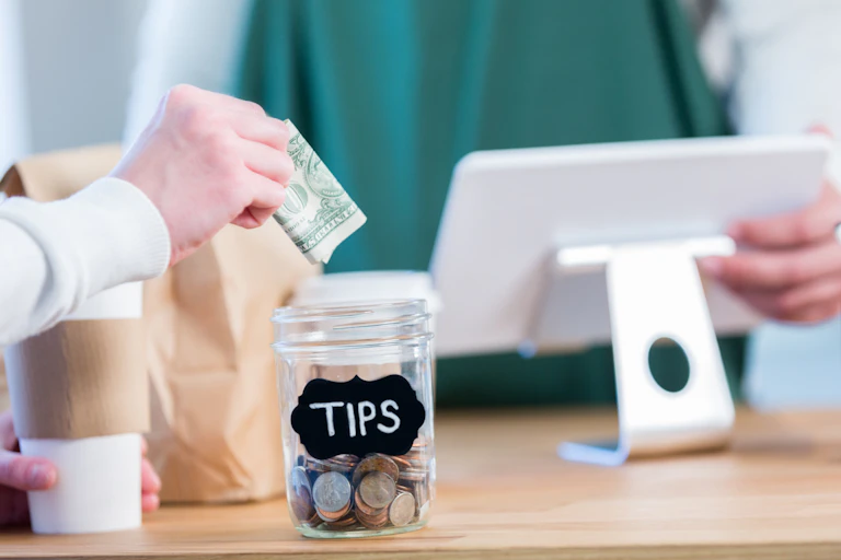 A white hand holds some dollar bills over a jar filled with change that has the word tips written in white letters on a black label, on a table with a white small screen and a coffee cup
