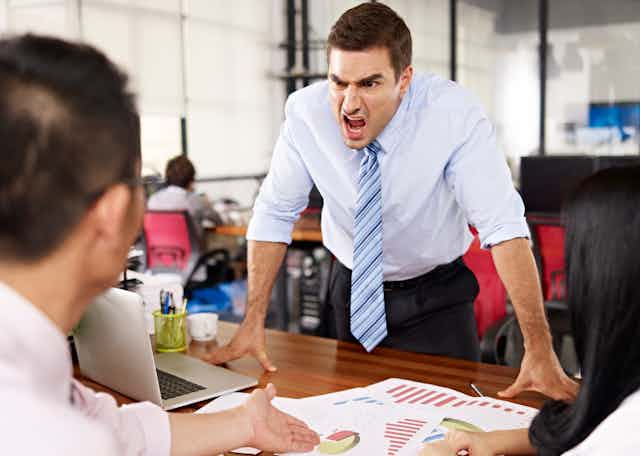 A white man appears to be angry and shouting while standing and leaning with both hands over a desk while another white employee, back turned, gestures to some spreadsheets on the table