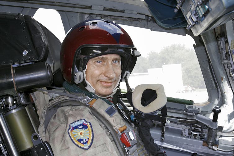 A man in a training suit wearing a helmet sits in the cockpit of a bomber jet.