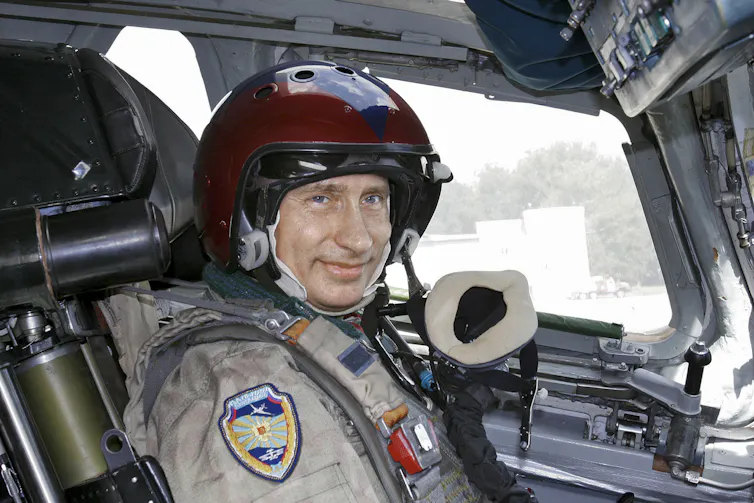 A man in a training suit wearing a helmet sits in the cockpit of a bomber jet.