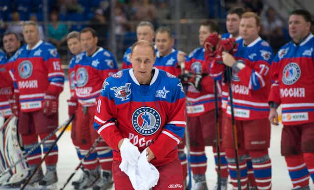 A balding man is seen in a red and blue hockey jersey on the ice with hockey players standing behind him.