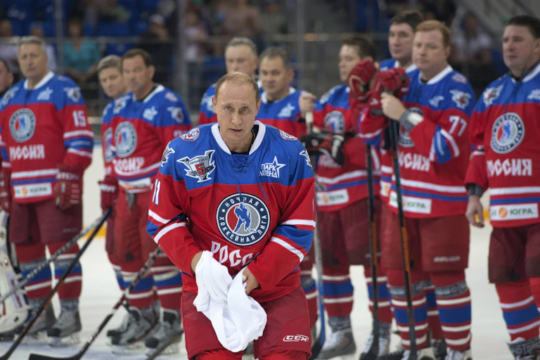 A balding man is seen in a red and blue hockey jersey on the ice with hockey players standing behind him.