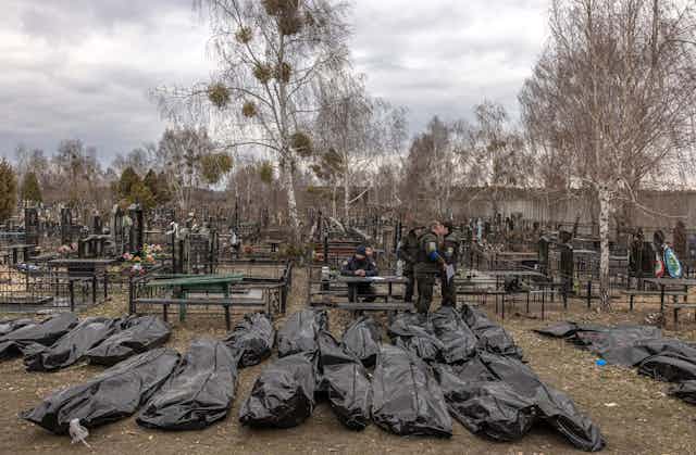 People in police uniforms walking among rows of dead bodies in black plastic bags in a cemetery in Bucha, Ukraine.