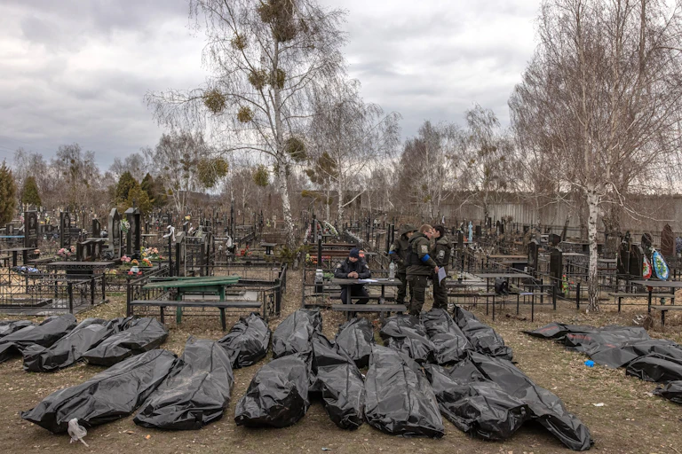 People in police uniforms walking among rows of dead bodies in black plastic bags in a cemetery in Bucha, Ukraine.