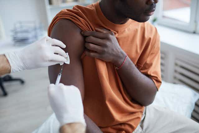 African Australian man in an orange tshirt raises his sleeve to get vaccinated.