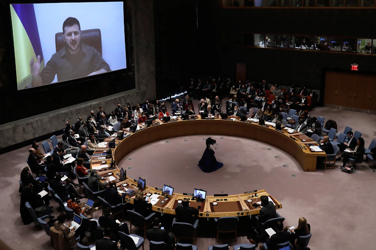 A man appears on a video screen addresses men and women sitting around a horseshoe shaped table filled with computers screens.