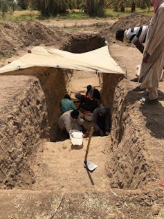 a tarp shades people working in a rectangular trench cut out of sandy dirt