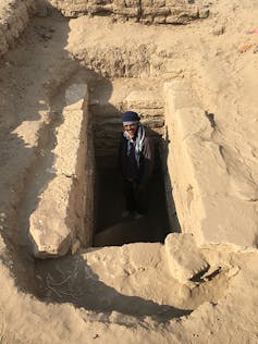 Man stands in deep rectangular hole cut from dusty dirt