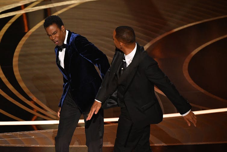 Man in tuxedo slaps another man in a tuxedo on stage.