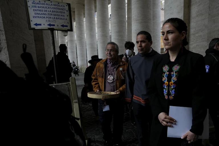 A woman in a black shirt holding a document stands in front of two men in St. Peter's Square at the Vatican.