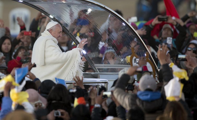 Wearing a poncho, Pope Francis waves from the popemobile as a large crowd of people surround him.
