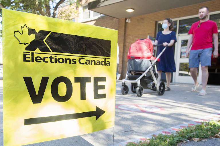 A yellow sign showing where to vote is in the foreground as a couple pushing a baby carriage leave a voting station.