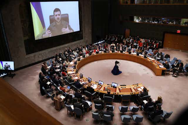 A man with dark hair on a screen with a Ukrainian flag behind him talks to a meeting where people are sitting in an almost-complete circle of desks.