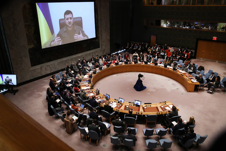A man with dark hair on a screen with a Ukrainian flag behind him talks to a meeting where people are sitting in an almost-complete circle of desks.