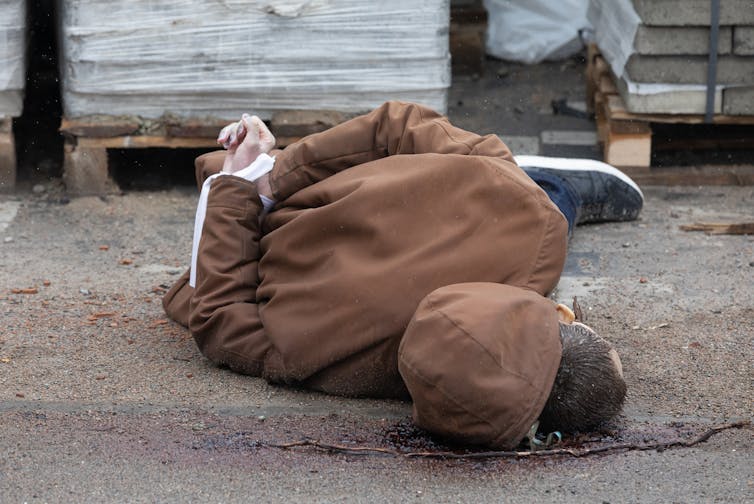 A dead body on the ground, dressed in a brown jacket with hands tied behind their back.