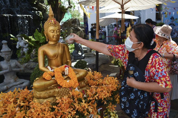 A woman wearing a face mask pouring water over a bronze-colored Buddha statue that is decorated with flowers.