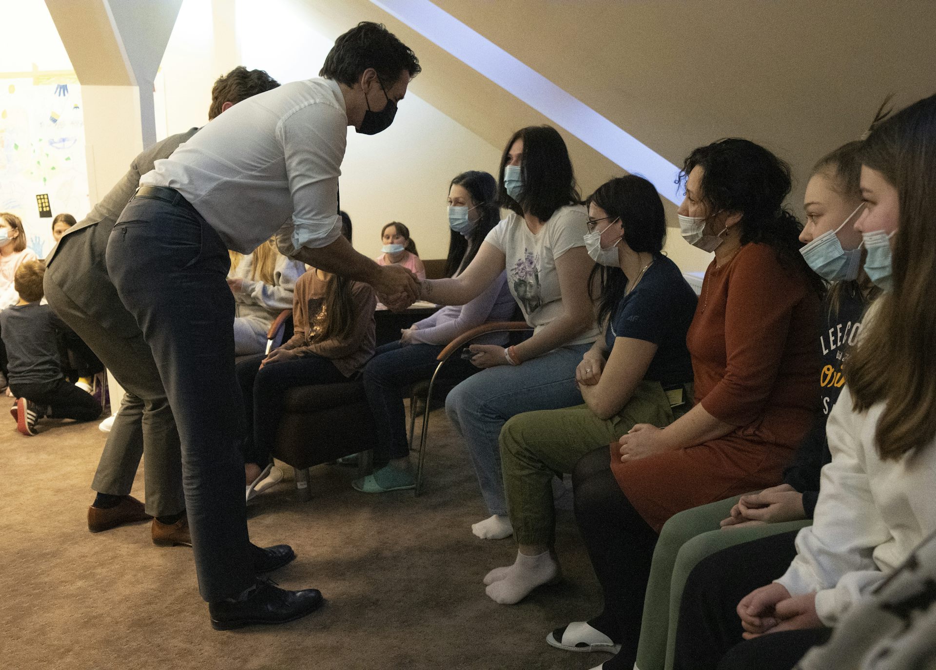 A dark-haired man wearing a mask shakes hands and talks to women and children.