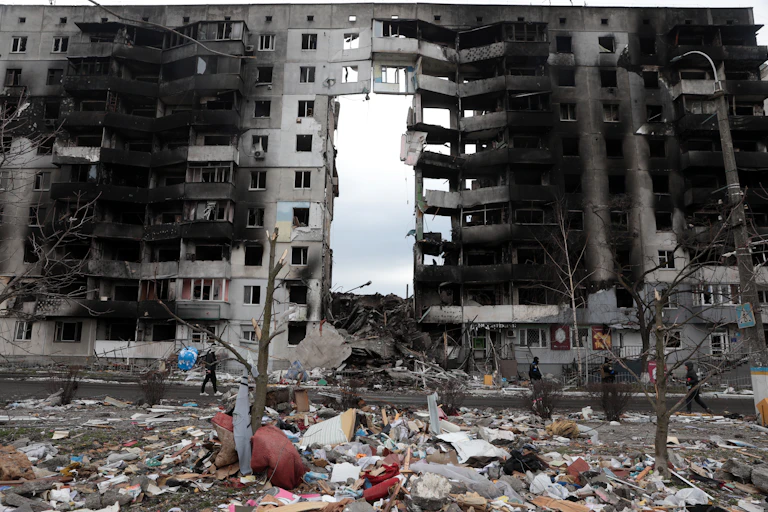 A destroyed apartment building, blackened by fire and with a huge hole in the middle. In front of it is rubble.
