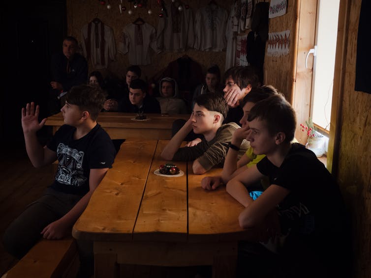 A group of teens in a dark room, sitting at a table, listening to someone speak.