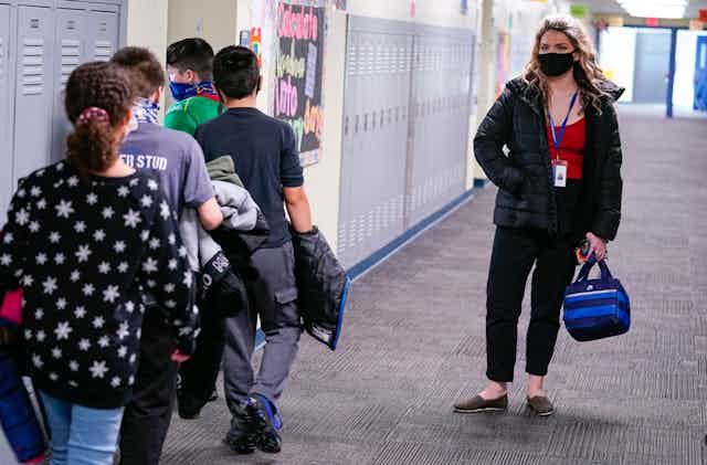 An adult wearing a jacket and carrying a lunch bag watches students file into a classroom