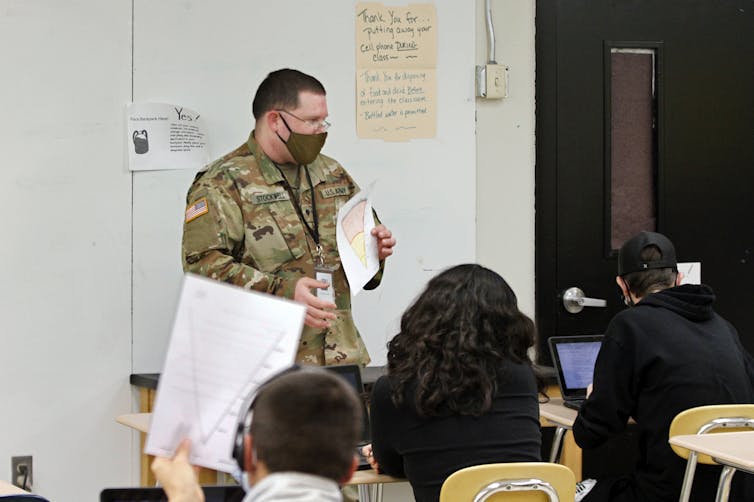 A person in a camouflage military uniform holds a paper in front of a class full of students