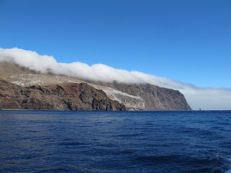 A photo of a large island from the sea with clouds spilling over.
