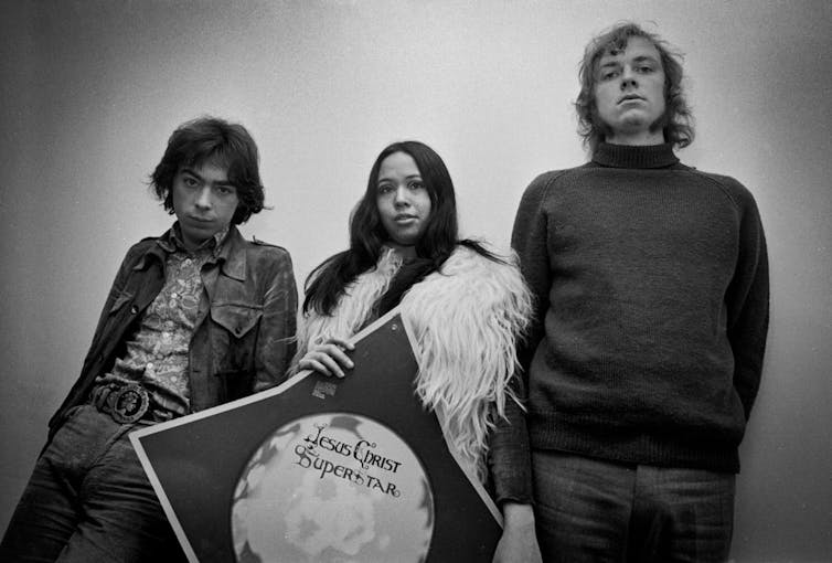 Two men stand on either side of a woman holding a sign that says 'Jesus Christ Superstar.'