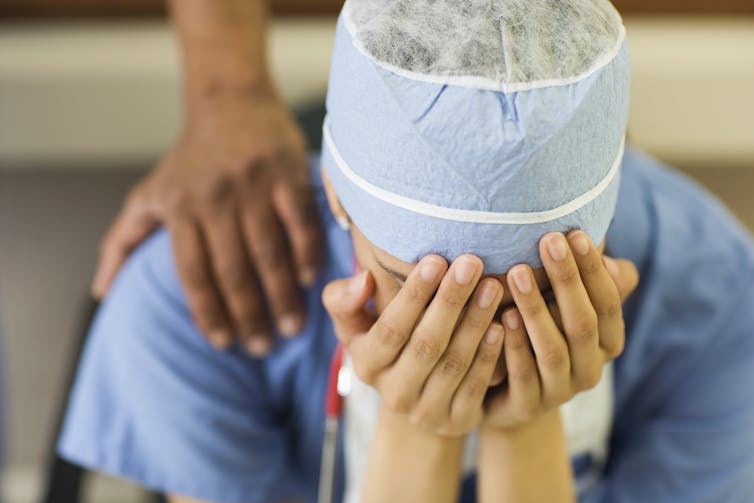 Person in scrubs with head in hands. A second person has their hand on their shoulders.