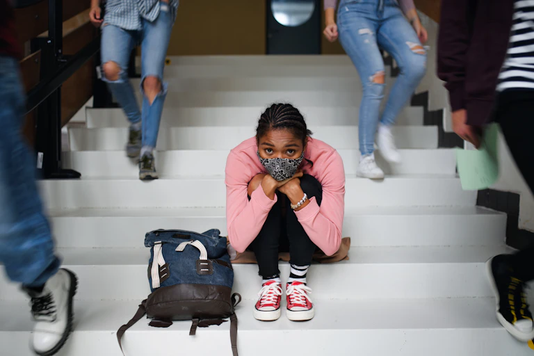 A young woman with face mask sitting on floor, with people walking around her.