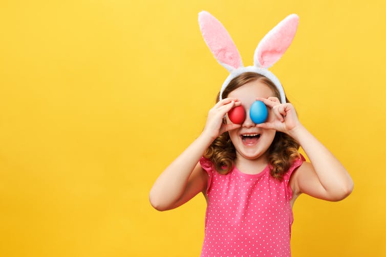 Against a bright yellow background, a young girl wearing bunny ears laughs and holds up two colourful eggs to her eyes.
