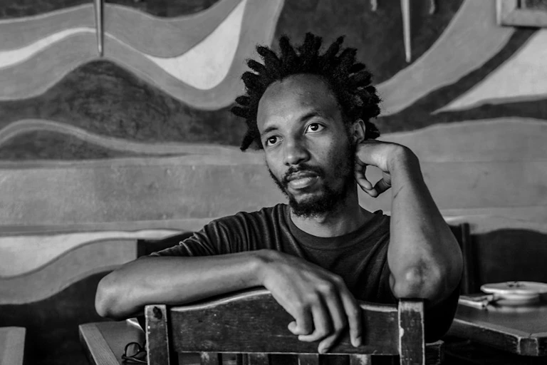A young man with his hair in spiky dreadlocks sits the other way round in a chair, his arm leaning against the back of it, another hand resting on his face. He looks into the distance without smiling.