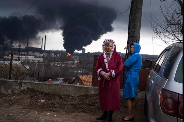 Deux femmes se tiennent à côté d'une voiture, avec un nuage de fumée noire en arrière-plan