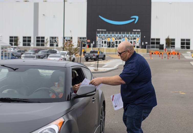 Man handing flier to the driver of a car. In the background stands a massive building with the Amazon logo.