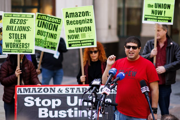 Man speaks into microphones while people holding protest signs that say 'Amazon Recognize the Union Now' stand behind him