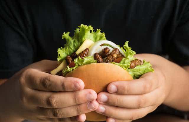 A close up of two hands holding a burger brimming with insects.