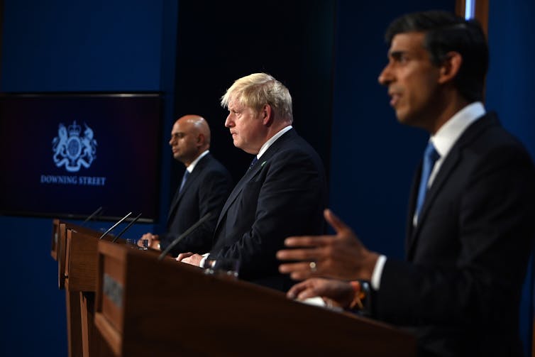 Side view of Rishi Sunak, Boris Johnson and Sajid Javid speaking behind podiums at a press conference