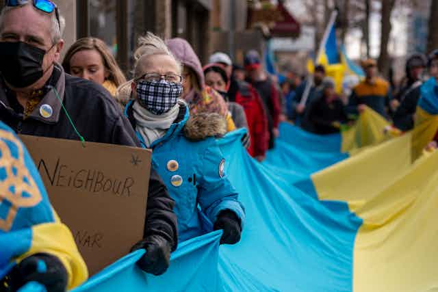 A line of people, some wearing masks, carry a giant blue-and-yellow flag of Ukraine.