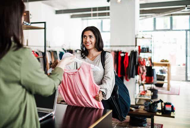 A smiling woman in a clothing store hands a dress to a cashier.