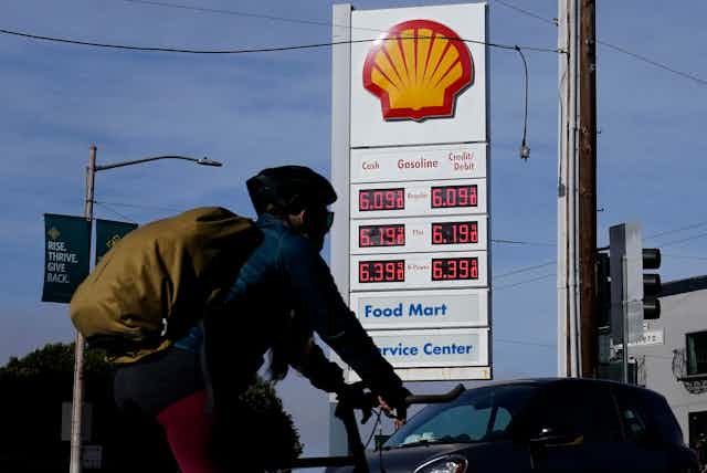 A man on a bike wearing a helmet and backpack bikes past a gas station sign with a price of 6.09 a gallon and higher