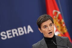 A dark-haired woman in a turtleneck and suit jacket speaks