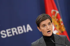 A dark-haired woman in a turtleneck and suit jacket speaks