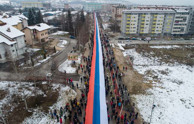 people march through a snowy city carrying a giant red, white and blue serbian flag