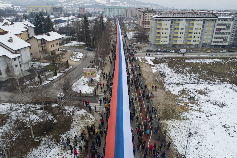people march through a snowy city carrying a giant red, white and blue serbian flag